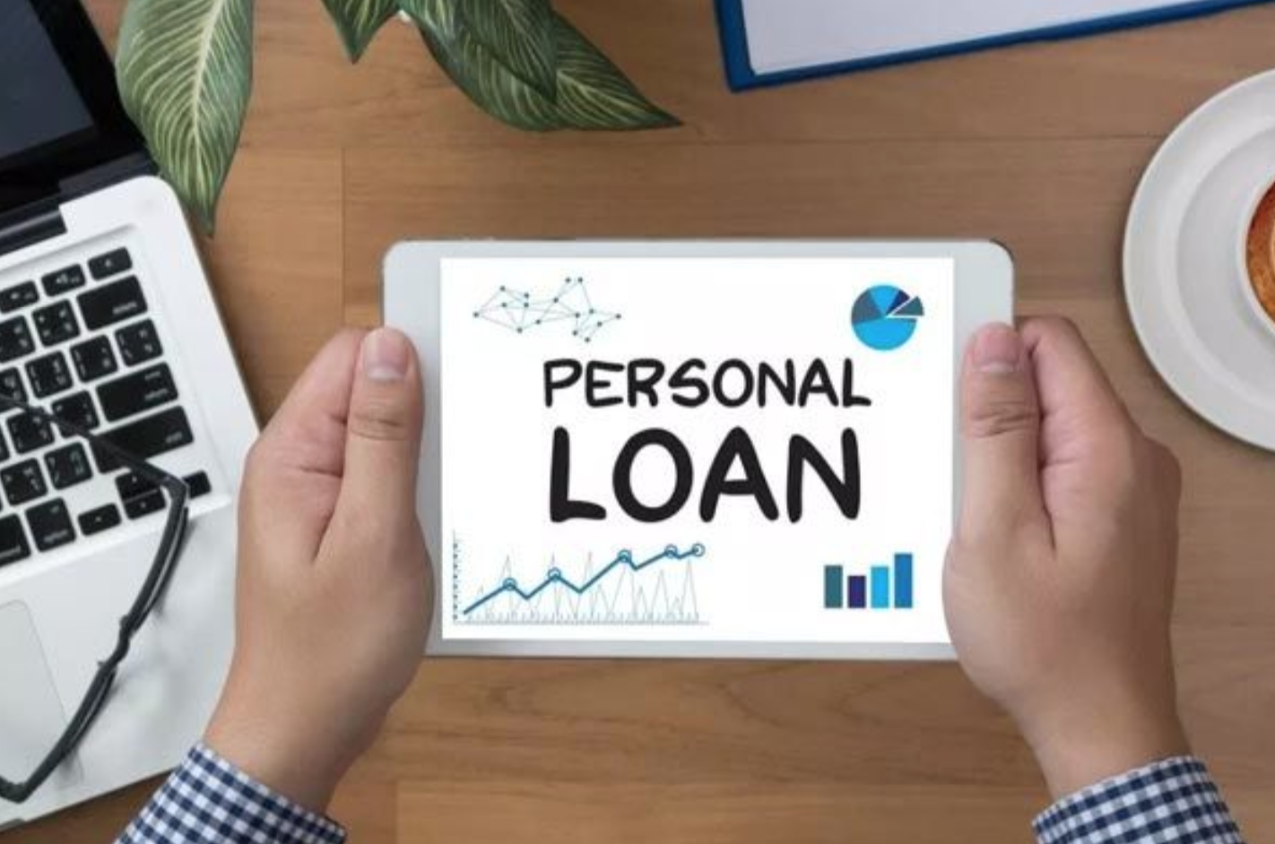  A person is holding a tablet that has a 'Personal Loan' graphic on the screen, with a laptop and cup of coffee on the table.